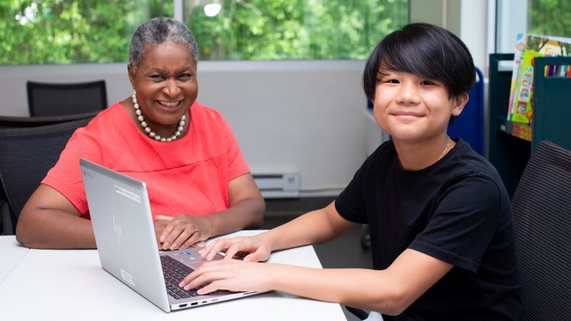 an adult sits next to a child, who is typing on a laptop