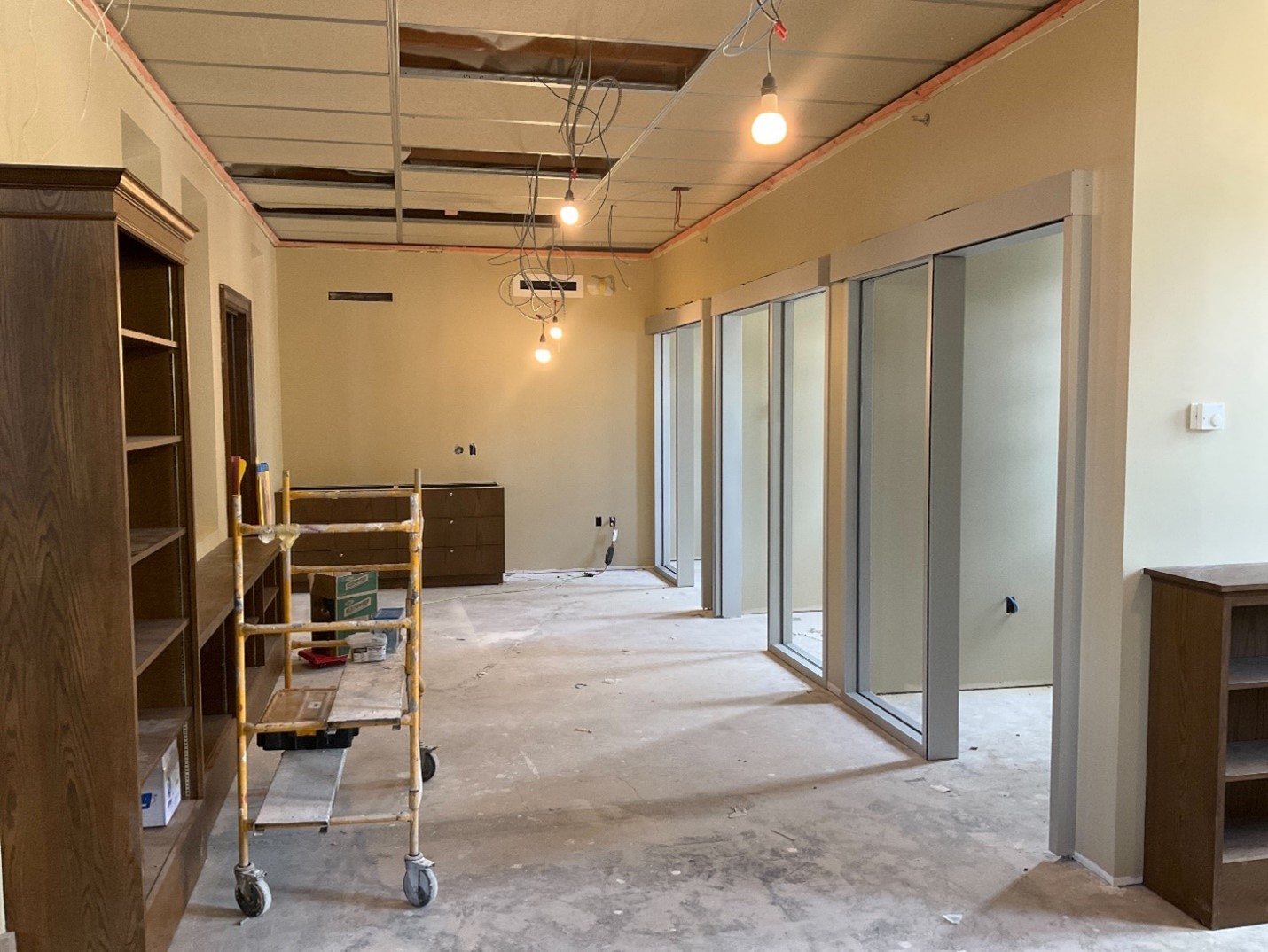 a sumner library room under construction with empty shelves and bare lightbulbs hanging from the ceiling. entrance to three meeting rooms without doors on the right side.
